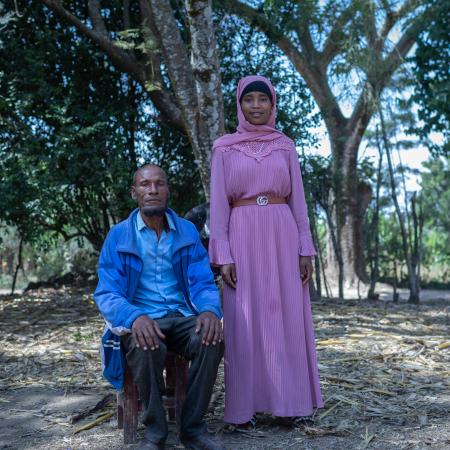 A man sits on a stool and his teenage daughter stands next to him. They are outdoors with many trees behind them.
