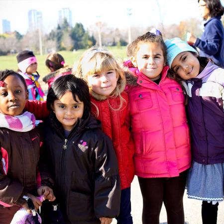 A diverse group of children stand shoulder to shoulder outside on a chilly but sunny day, smiling at the camera.