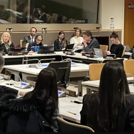 A group of delegates sit around a conference table with nameplates, engaged in discussion. The setting is formal, suggesting a diplomatic meeting.