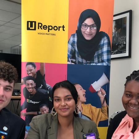 Three young people holding up cellphones in their hands and smiling. Behind them is a standee with a poster that shows the U-Report logo.