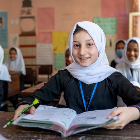 A girl with her textbook open smiles at the camera while sitting in a classroom in Kabul, Afghanistan.