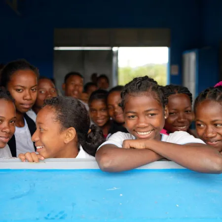 Un groupe de plus de dix enfants souriants regardent dehors depuis la fenêtre d’une salle de classe. 