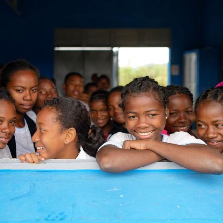 A group of more than ten smiling children look outdoors from a classroom window.