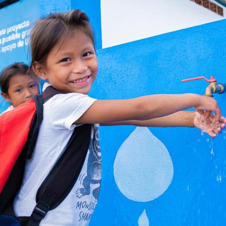 A girl smiles at the camera while washing her hands at a UNICEF tap.    