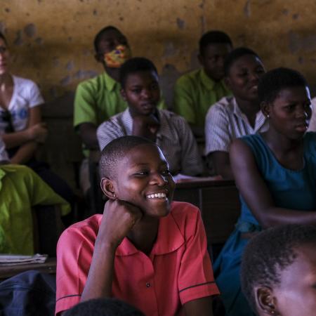 A classroom in Ghana with students sitting at desk. In the centre of the photo is a girl in a pink uniform smiling while looking beyond the camera. 