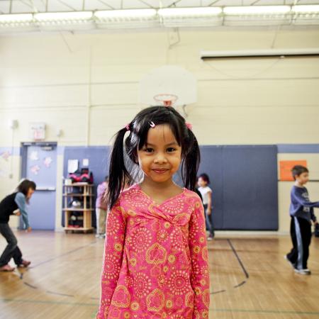 A young girl stands smiling at the camera on a basketball court, while her classmates play in the background. 