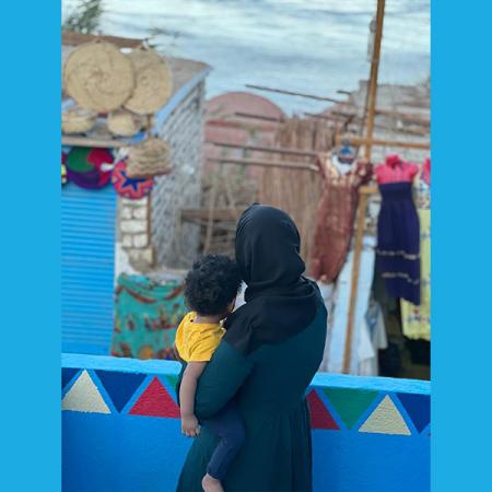 A woman looks out from a balcony, her back is towards the camera and she is holding a child in her arms. 