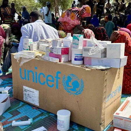 A UNICEF box covered with medical supplies sits on a colorful mat. In the background, people in vibrant clothing gather, conveying a sense of community.