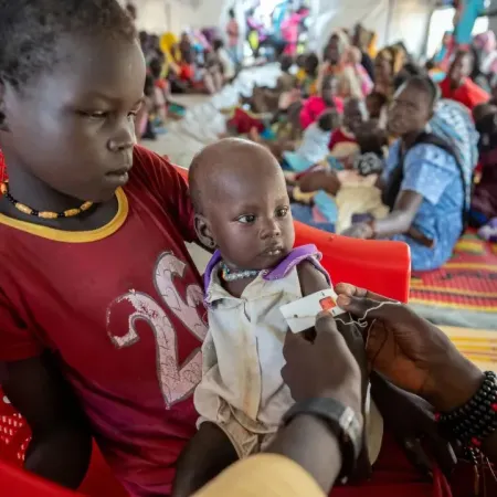 A young child sits in a red chair, holding an infant being assessed for malnutrition in a crowded room. The scene is serious and focused on well-being.