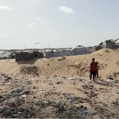 Two children stand in a sandy field with garbage around overlooking tents