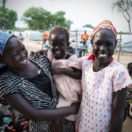 Two women smile, while looking at the camera, in between them is a child in a dress also smiling, the child is being held by the woman on the left. There are white tents behind them. This photo was taken in South Sudan in 2015. 