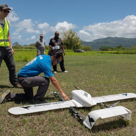 A man in a UNICEF t-shirt bends down to check on a small white drone in a field in Vanuatu.