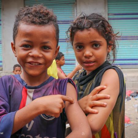 Two children on a street in Yemen, show off their arms with their sleeves rolled up, poking their arm to mimic a vaccine shot with their hands. 