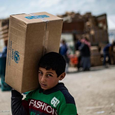 A child in a refugee camp carries a box of UNICEF supplies as he walks. This photo was taken in Syria in 2019. 