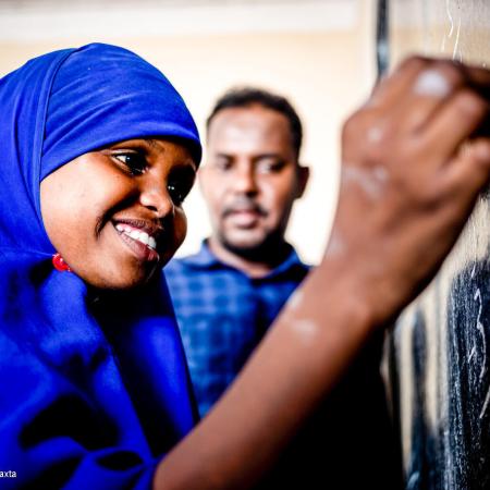 A girl wearing a blue uniform smiles as she writes on a chalk board as her teacher looks on in the background. This photo was taken in Somalia in 2021. 
