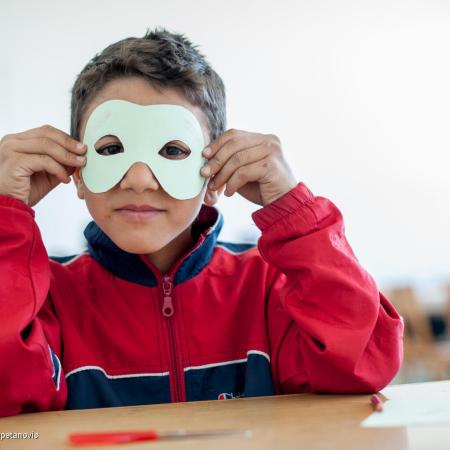 A child wearing a red jacket sits in a classroom desk. He is holding up a white paper mask with eyes cut out and holding it to his eyes. This photo was taken in Bosnia and Herzegovina in 2013.