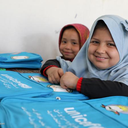 Two girls sitting at a desk with UNICEF bagpacks in front of them, both are looking at the camera and smiling. This photo was taken in Afghanistan in 2021. 