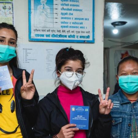 Three female health workers give the 'v for vaccinated' sign outside a clinic in Nepal.