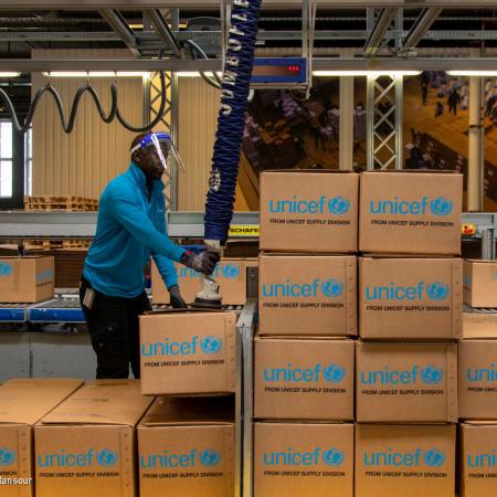 A man uses a robotic arm in a warehouse. There are boxes with UNICEF labels stacked in front of him. This photo was taken in Denmark in 2021. 