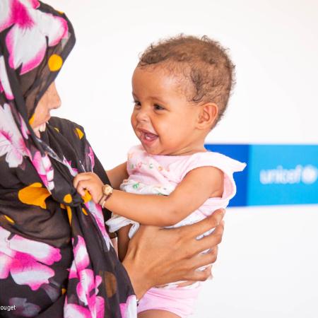 A mother holds her daughter, the mother is wearing a floral-patterned scarf and the child is wearing a pink dress. The baby and mother are both smiling. This photo was taken in Mauritania in 2021.