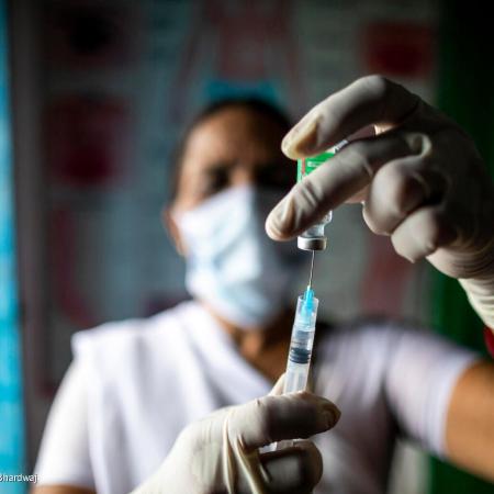 A health worker uses a syringe to extract a vaccine from its vial. He is wearing gloves. The syringe and vaccine vial and his hands are in focus and he is in the background out of focus. This photo was taken in India in 2021. 