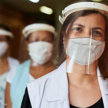 Three women stand in a line, the first one is in focus. They are all wearing masks and PPE headgear. This photo was taken in Brazil in 2021.