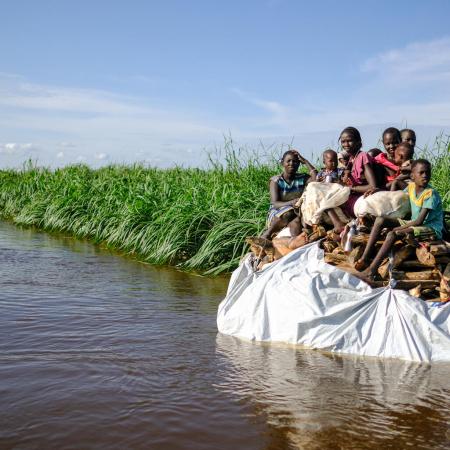 A group of children sit on top of a tarp of belongings on the sides of a flooded river bank.