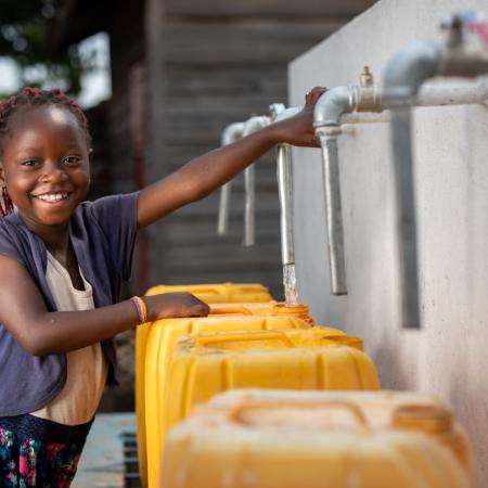 A girl smiles at the camera while filling a yellow water jug at an outdoor tap.