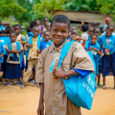 A child with a blue UNICEF bag pack slung on one side smiles at the camera. He is wearing a brown uniform, behind him out of focus are many children also wearing bag packs and brown uniforms.  This photo was taken in the Congo in the province of Lékoumou. .  