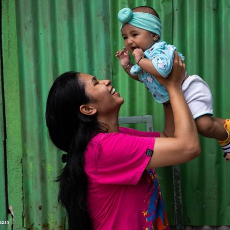 A woman wearing a pink tshirt holds her infant above her head, smiling. The infant is also smiling. 