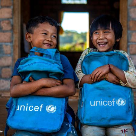 Two children holding UNICEF backpacks sit at the stoop of a classroom, smiling. 