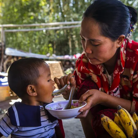 A woman in a red sweater feeds a young child with a spoon from a bowl; bananas and baskets are nearby, and trees are visible in the background.