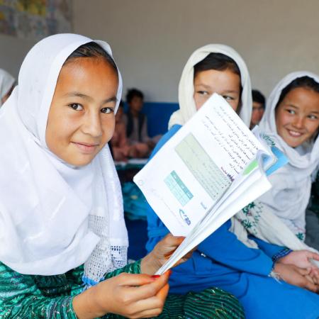 At a classroom in Afghanistan, a girl wearing a white headscarf holds open a book and smiles at the camera.
