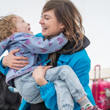 A woman wearing a UNICEF jakcet carries a child in her arms, both the woman and child are laughing. They are outdoors and both are wearing jackets.