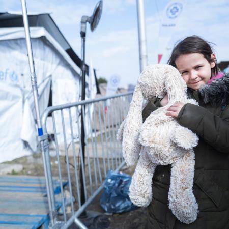 A child holds a bunny as she looks at the camera with a small smile. She is wearing a jacket and standing outdoors, in a line, behind her are white UNICEF tents. This photo was taken at the Ukrainian border in 2022. 