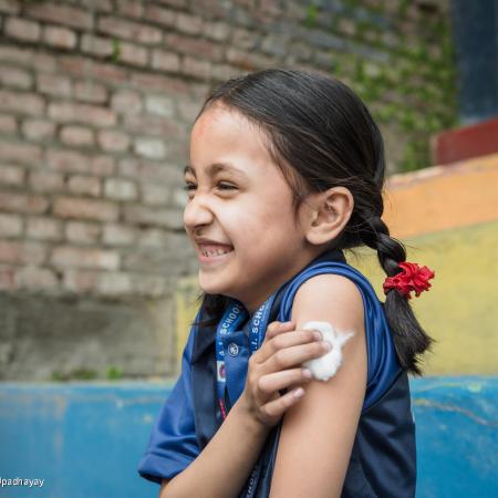 A girl holds a piece of cotton up to her recently vaccinated arm. She is smiling.  This photo was taken in Nepal in 2022. 
