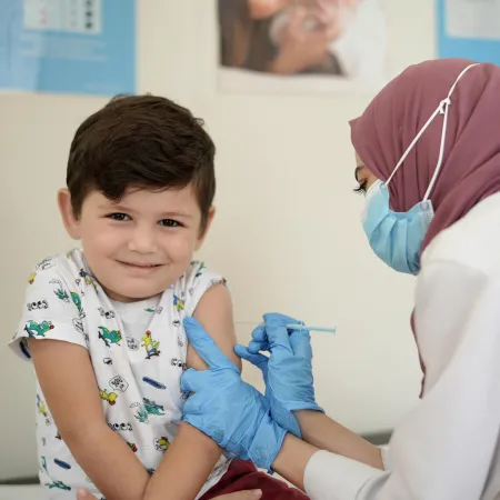 A child looks at the camera with a shy smile, a health care worker in a mask and medical gloves gives him a shot in the arm. The setting looks like a clinic. 