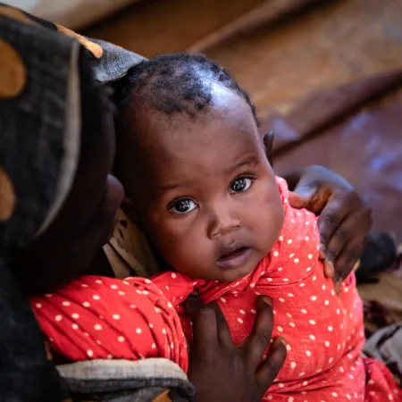 A child wrapped in a red polka dotted blanket looks up to the camera. he is held by his mother whose face is cast down.