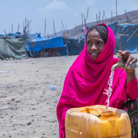 A girl in a bright pink scarf in front of yellow jerry can which has water pouring in it from a single tap. Tents can be seen in the background, looking like she is in a camp of sorts. The girl is looking away from the camera and is smiling. 