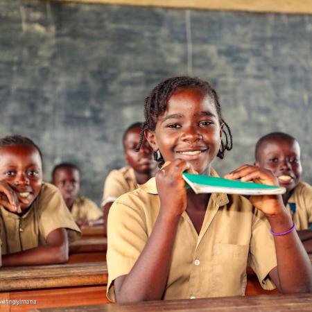 A girl sits in a classroom smiling. She is holding up a green notebook. Behind her are other children in brown uniforms, also smiling. This photo was taken in the Congo in 2022. 