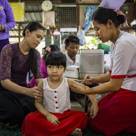 A girl sitting cross legged, looking very calm while getting an injection by a nurse, another woman is next to the child, looking at her. 