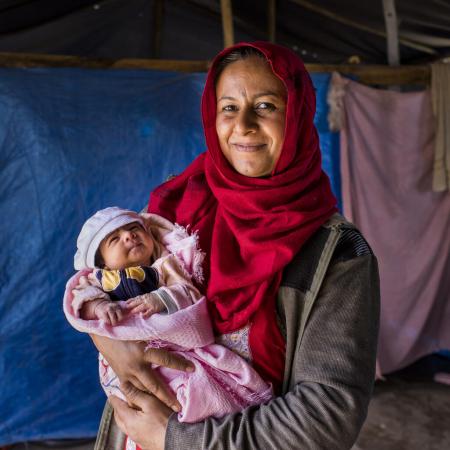 A mother in front of a tent holding a newborn in pink blanket. 