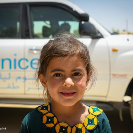 An Afghan girl looks at the camera with a shy smile, behind her is a white vehicle with the words UNICEF written on it. 