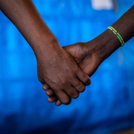 Hands being held, the hand on the left is that of a man and the one on the right of a girl. The girl is wearing a green beaded bracelet. The photo is cropped till the forearms, the background is that of a blue tarp.