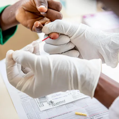 A close up of a child's hand being pricked by a medical professional's hands who is wearing white latex gloves.