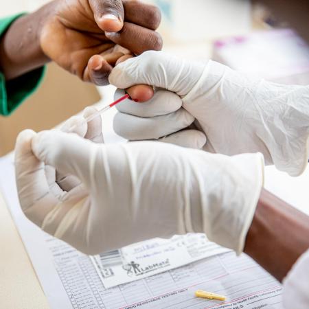 A close up of a child's hand being pricked by a medical professional's hands who is wearing white latex gloves. 