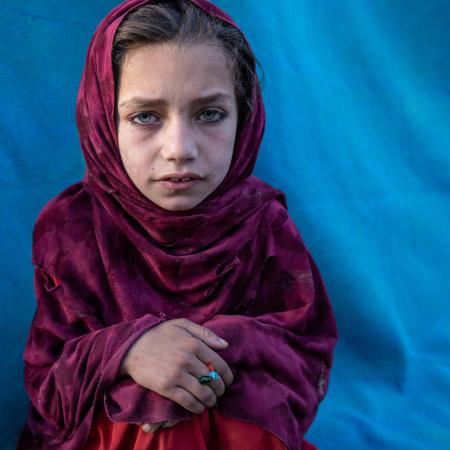 A girl wearing a magenta headscarf while sitting, looking at the camera, with her hands folded on her knees. There is a vivid blue tent behind her