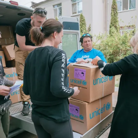 A van with the back door open and a several boxes with the words UNICEF written on them. Two women stand in front of the boxes looking at them. One man is unloading the boxes and two other men are standing to the side.