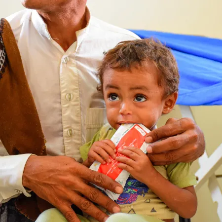 A man holds a child who is eating from a red packet of Plumpy'Nut. This photo was taken in Yemen in 2022.