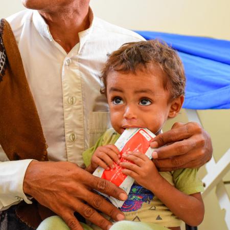 A man holds a child who is eating from a red packet of Plumpy'Nut. This photo was taken in Yemen in 2022.